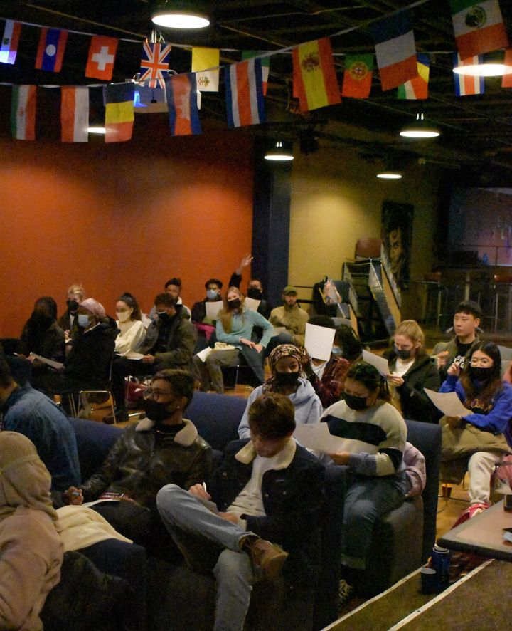 A large group of students sit in a dark room eating food and playing games